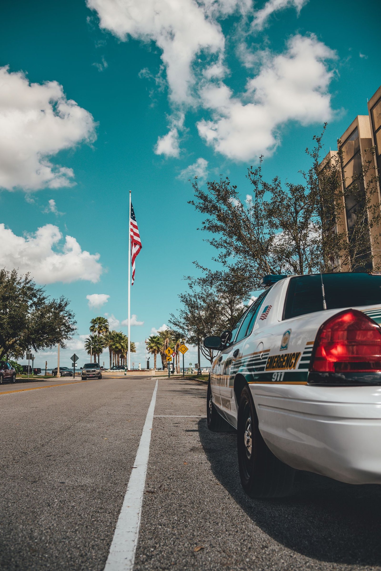A police car parked by a road under a clear sky with an American flag and trees in the background.