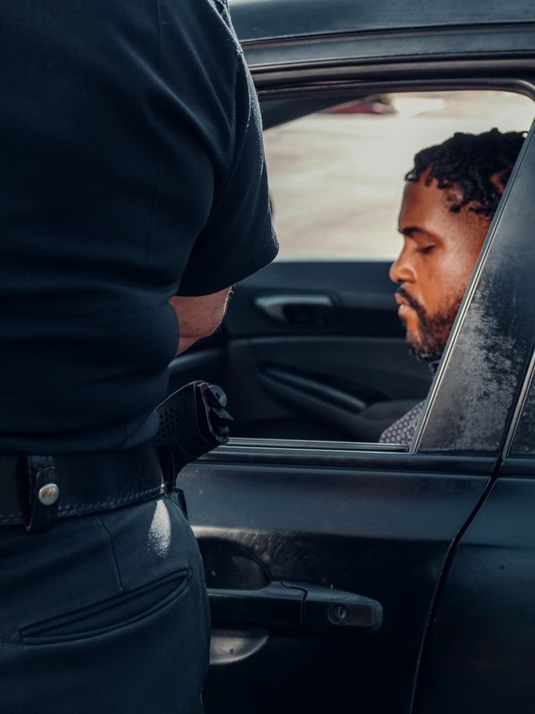 Police officer inspecting a vehicle with driver seated inside, sunny day.